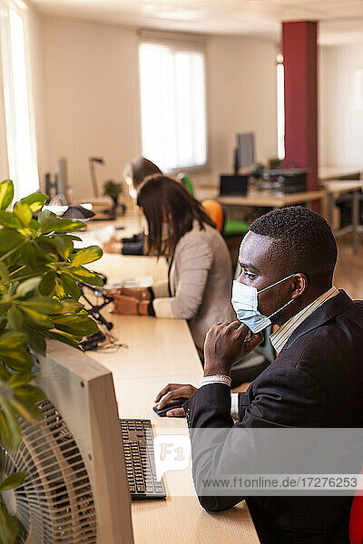 Männlicher Unternehmer mit Schutzmaske bei der Arbeit am Computer in einem Coworking-Büro während des Ausbruchs des Coronavirus
