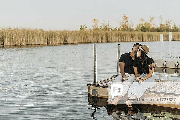 Man with wineglass kissing woman while sitting on pier