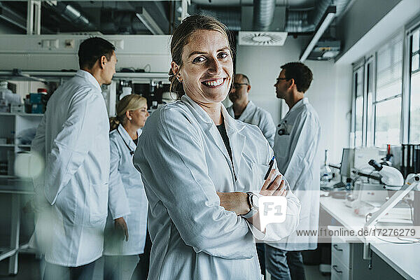 Lächelnde Frau mit verschränkten Armen  die mit einem Mitarbeiter im Hintergrund in einem Labor steht