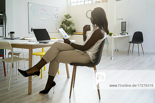 Woman with coffee cup reading paper while sitting on chair at office