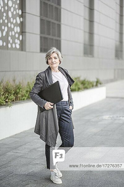 Confident businesswoman holding laptop while standing with hand in pocket against building