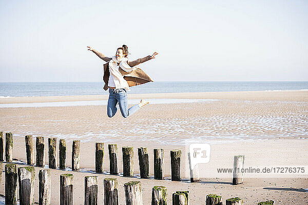 Happy young woman with arms outstretched jumping from wooden posts at beach during sunny day