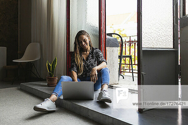 Lächelnde kreative Geschäftsfrau  die am Laptop arbeitet  während sie an der Tür im Büro sitzt