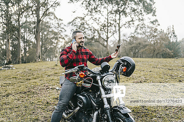 Männlicher Motorradfahrer  der auf seinem Motorrad im Wald sitzt und mit seinem Smartphone telefoniert