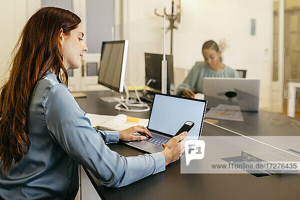 Geschäftsfrau mit Smartphone und Laptop am Schreibtisch sitzend