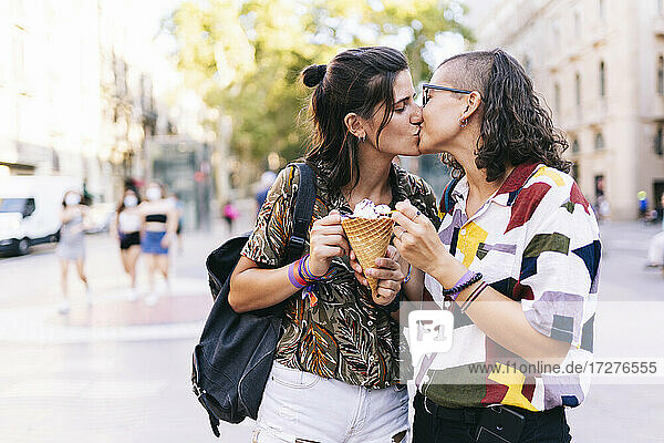 Young lesbian couple holding ice cream while kissing on street in city