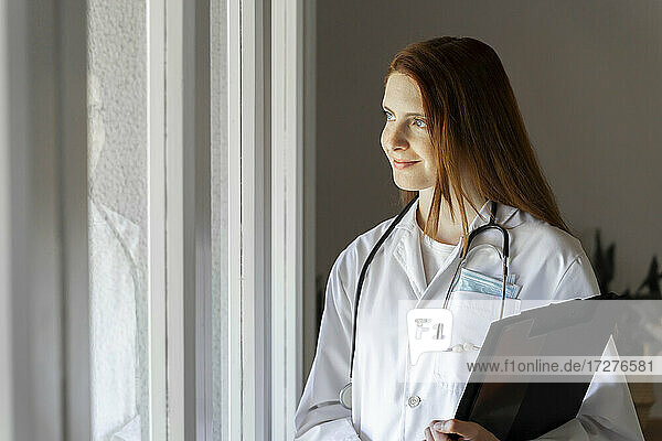 Nachdenkliche junge Ärztin lächelt  während sie durch das Fenster im Büro zu Hause schaut