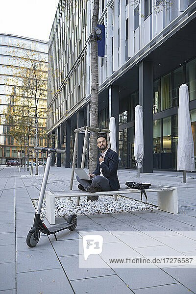 Smiling businessman using laptop by electric push scooter on bench in city