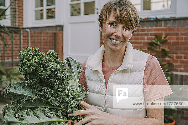 Lächelnde erwachsene Frau  die ein Grünkohlblatt hält  während sie im Hinterhof steht