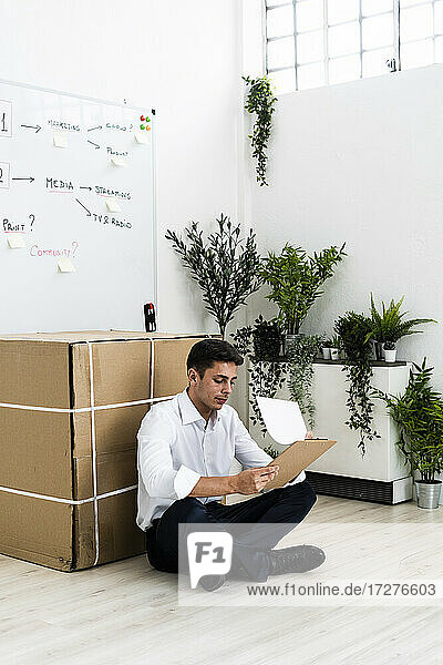 Businessman looking at clipboard while sitting on floor against cardboard box in office