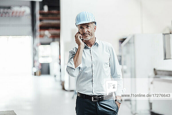 Männlicher Ingenieur schaut weg  während er in einer Fabrik mit einem Mobiltelefon spricht