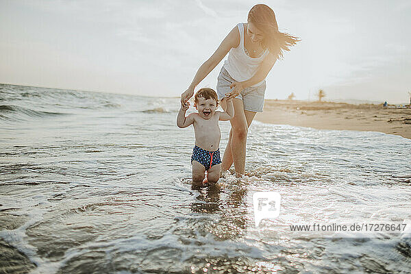 Mother and son enjoying in water at beach on sunny day during sunset