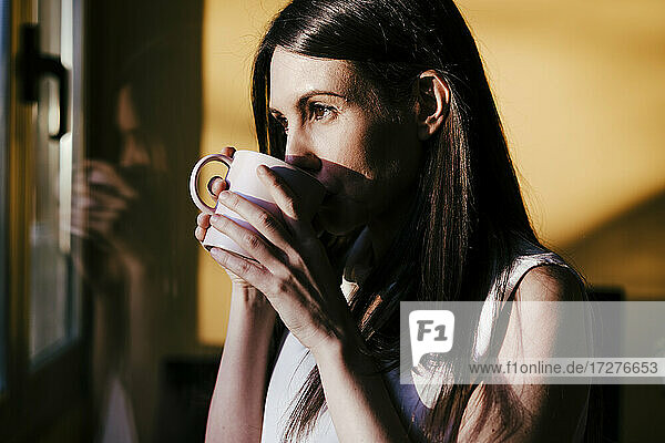 Frau schaut durch das Fenster und trinkt Kaffee im Stehen zu Hause