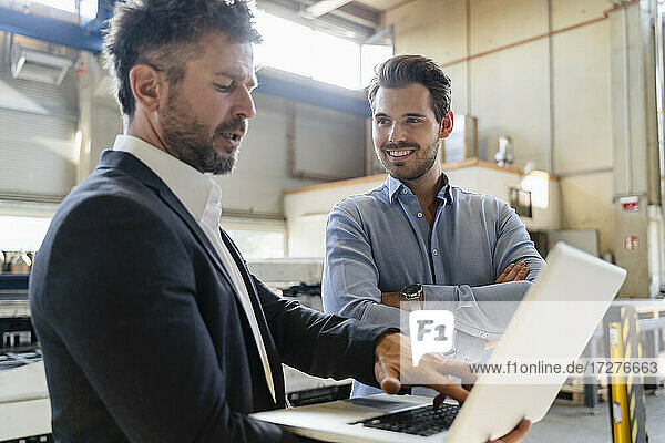 Ein lächelnder Kollege steht neben einem Geschäftsmann  der in einer Fabrik einen Laptop benutzt.