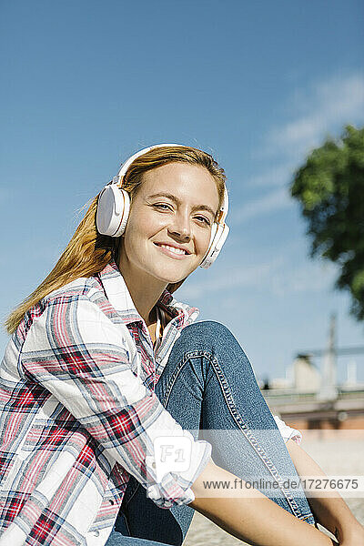 Lächelnde Frau  die mit Kopfhörern Musik hört  sitzt auf einem Fußweg an einem sonnigen Tag