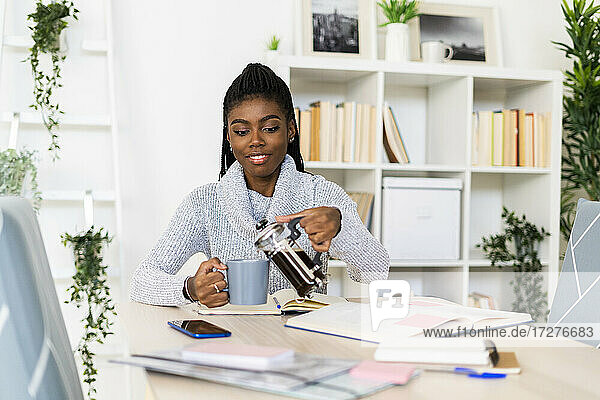 Lächelnde Frau gießt Kaffee in eine Tasse  während sie zu Hause sitzt