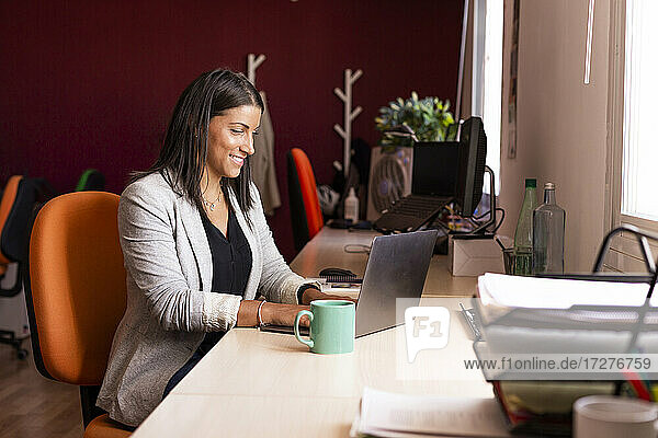Lächelnde Geschäftsfrau mit Laptop am Schreibtisch in einem Coworking-Büro
