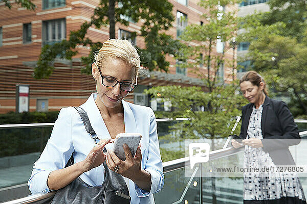Woman using mobile phone while colleague standing in background at city