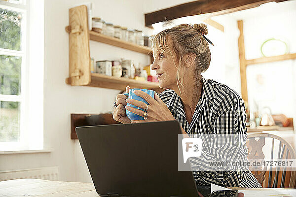 Aktive ältere Frau  die Kaffee trinkt und wegschaut  sitzt mit Laptop in der Küche
