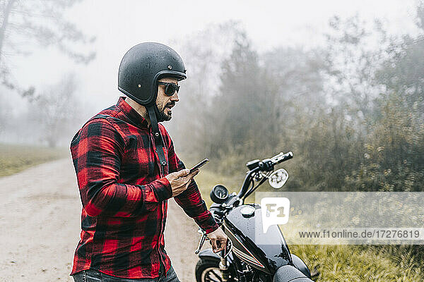 Männlicher Motorradfahrer  der über den Lautsprecher seines Smartphones spricht  während er bei nebligem Wetter neben seinem Motorrad steht