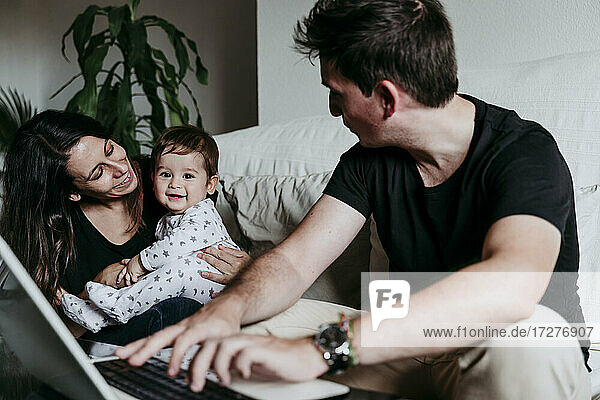 Smiling mother holding son while father working on laptop sitting at home