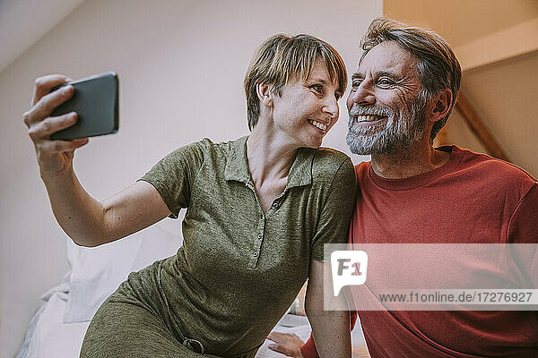 Smiling woman taking selfie while mature man sitting beside on bed at home