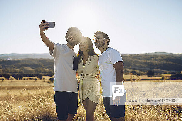 Smiling male and female friends taking selfie through smart phone while standing on field in summer