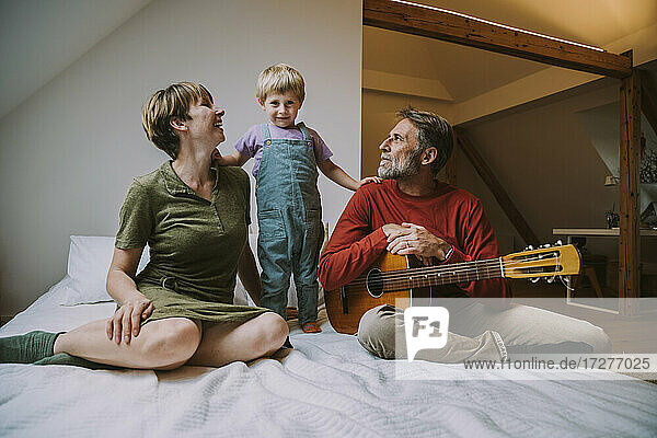 Son standing in between mother and father playing guitar sitting at home in bedroom