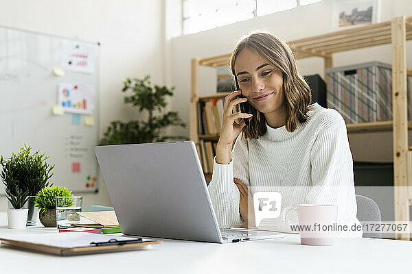 Geschäftsfrau  die mit einem Mobiltelefon spricht  während sie im Büro an einem Laptop arbeitet