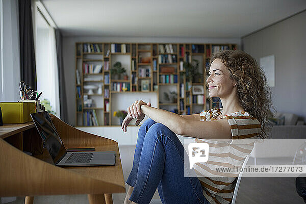 Lächelnde Frau  die wegschaut  während sie zu Hause auf einem Stuhl am Schreibtisch sitzt
