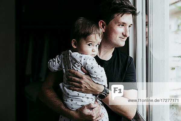 Smiling father carrying son while standing looking through window at home