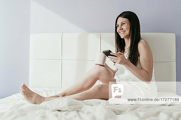 Lächelnde schöne Frau hält Smartphone und Kaffeetasse  während sie auf dem Bett zu Hause sitzt
