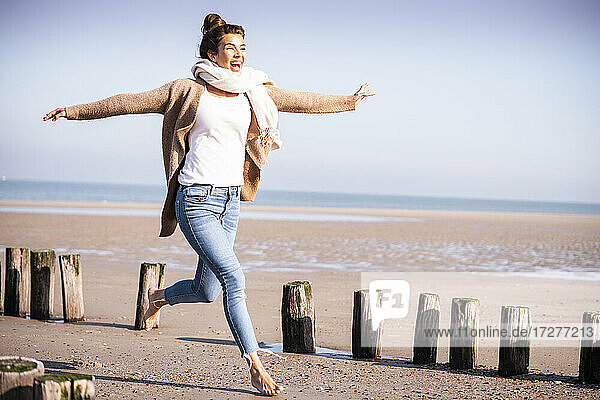Happy young woman with arms outstretched running amidst wooden posts at beach during sunny day