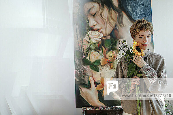 Nachdenkliche erwachsene Künstlerin  die eine Sonnenblume hält  während sie vor einem Kunstwerk im Atelier steht