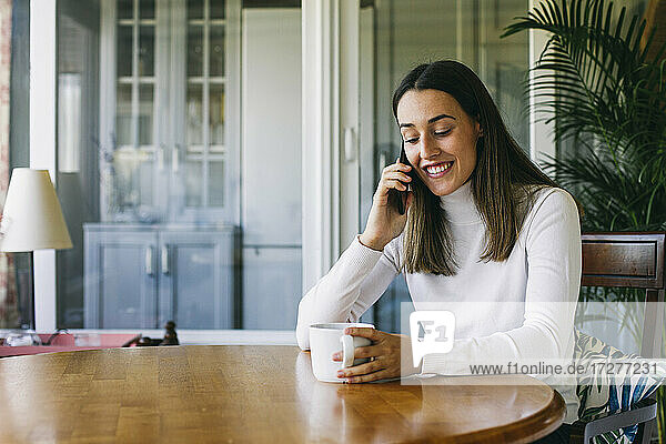 Lächelnde Frau mit Kaffeetasse  die mit ihrem Handy telefoniert  während sie zu Hause sitzt