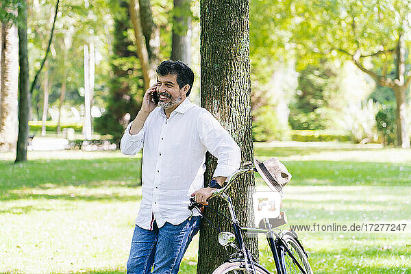 Glücklicher reifer Mann  der mit seinem Smartphone spricht  während er sich in einem öffentlichen Park an einen Baum lehnt