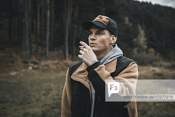 Young man looking away while smoking standing in forest during autumn