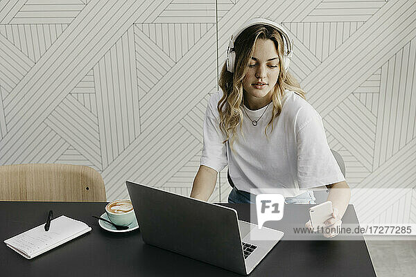 Junge Frau mit Kopfhörern  die an einem Laptop arbeitet und dabei ein Smartphone benutzt  während sie in einem Café sitzt