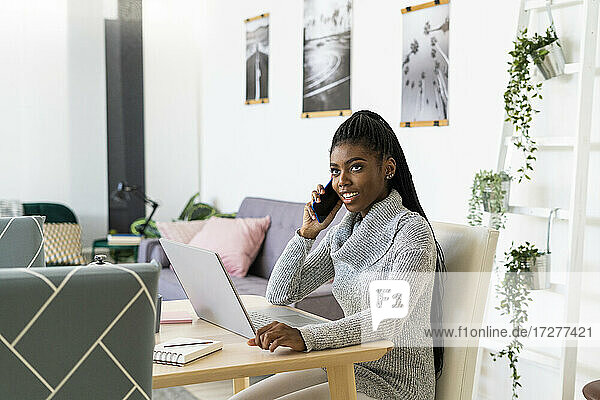 Lächelnde Studentin  die mit ihrem Smartphone spricht  während sie zu Hause am Laptop lernt
