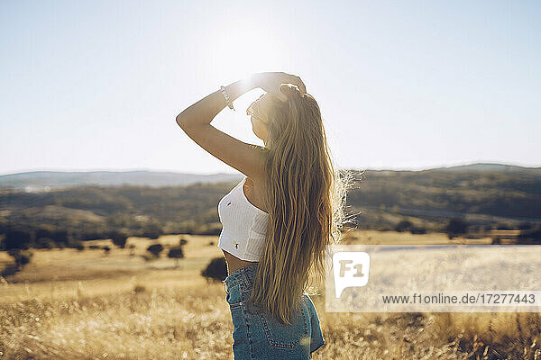 Junge Frau mit Hand in den Haaren steht auf einem Feld gegen den Himmel an einem sonnigen Tag