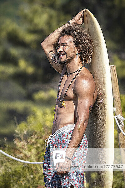 Lächelnder muskulöser junger Mann  der am Strand stehend ein Surfbrett hält