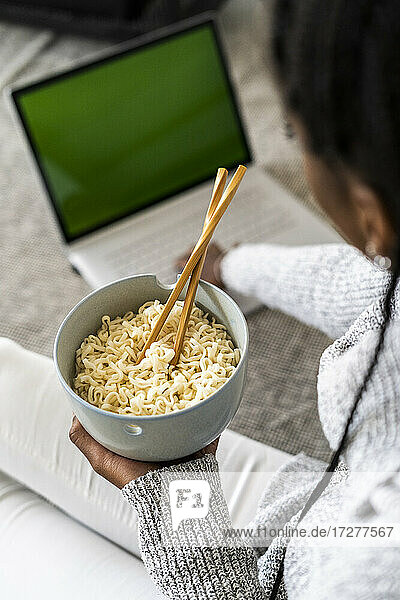 Frau hält Schüssel mit Nudeln  während sie einen Laptop zu Hause benutzt