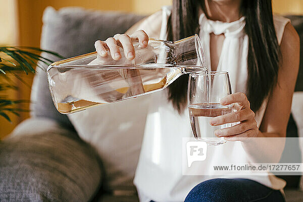 Frau gießt Wasser aus einem Glas in ein Trinkglas  während sie zu Hause sitzt