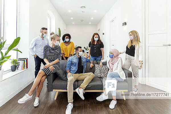 Männlicher und weiblicher Angestellter mit Gesichtsmaske sitzend und stehend am Sofa im Büro