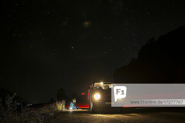 Weiblicher Camper sitzt vor einem Minivan mitten auf einer Landstraße in der Nacht