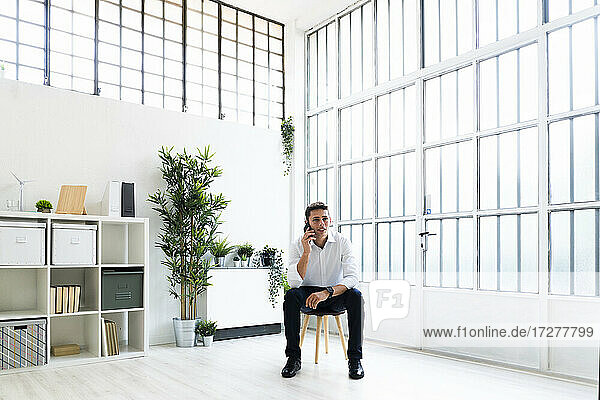 Young businessman talking through mobile phone while sitting on chair in bright creative office