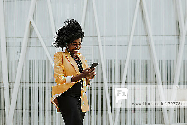 Lächelnde Unternehmerin  die ein Smartphone benutzt und über Kopfhörer Musik hört  vor einem Gebäude stehend