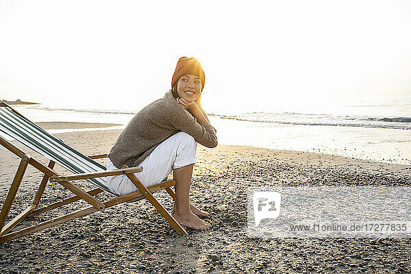 Lächelnde junge Frau  die auf einem Klappstuhl sitzt und bei Sonnenuntergang gegen den klaren Himmel auf den Strand blickt
