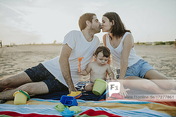 Ehemann und Ehefrau küssen sich  während ihr Sohn am Strand mit Spielzeug spielt
