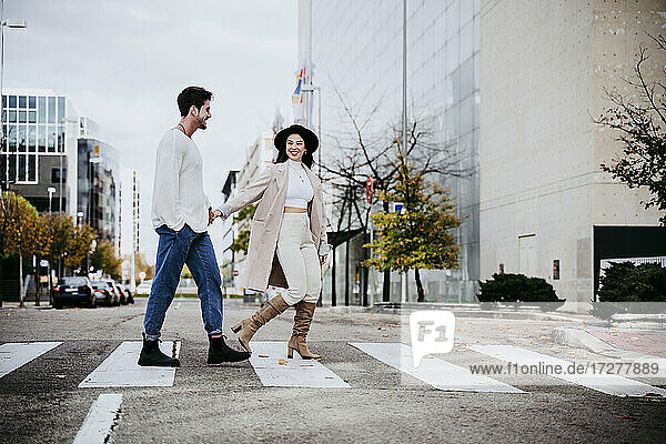 Lächelnde Frau hält Hände mit männlichem Partner beim Überqueren der Straße in der Stadt